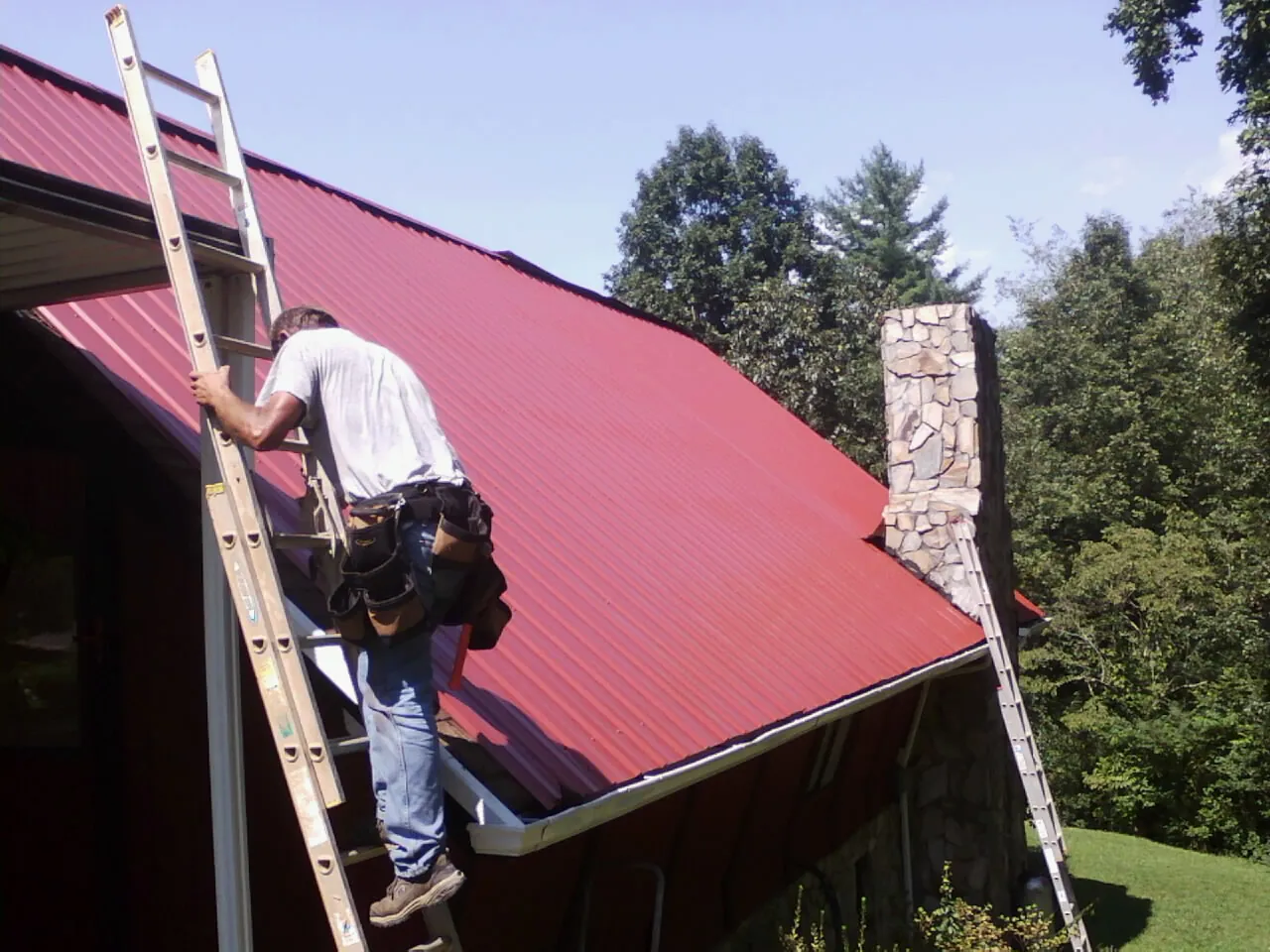 Roofer on a ladder beside a red metal roof and stone chimney — Soffit & Fascia Repair by Roofing Brothers in Bethel Island